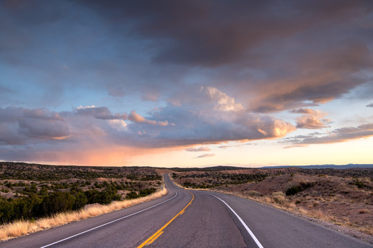 Highway curving into the distance through the landscape near Santa Fe,  New Mexico underneath a dramatic colorful sky at sunset