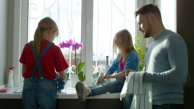 Medium shot of father and two little daughters washing dishes together and wiping them with cloth in kitchen