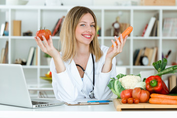 Beautiful young nutritionist looking at camera and holding fresh vegetables in the consultation.