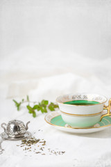 Royal Stafford Fine Bone China Tea Cup and Saucer with Loose Leaf Mint Tea, Spring of Fresh Mint in Background, and Tea Infuser on White Background