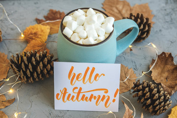 Blue mug with coffee, hot chocolate or cocoa with marshmallow on gray concrete background and lying dry leaves, cones, garland, sign Hello Autumn. Concept of warm cozy autumn. © tumana_net