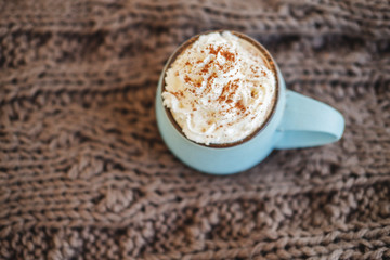 Mug of coffee, cocoa or hot chocolate with whipped cream and cinnamon on scarf. Pumpkin latte - cozy drink for cold autumn or winter.