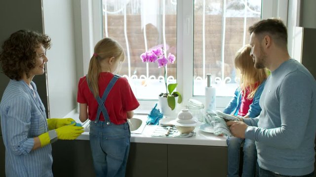 Medium shot of family with two daughters talking to each other while doing chores together in kitchen