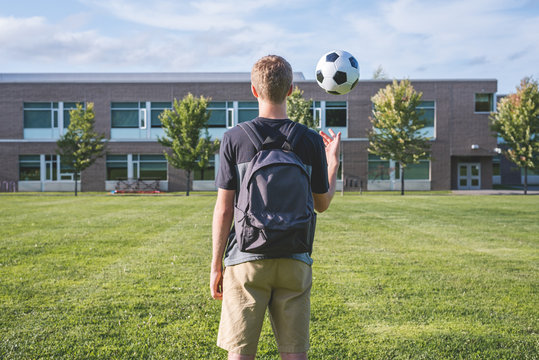 Teenager tossing a soccer ball up in the air as he stands on the sidelines of a soccer field.