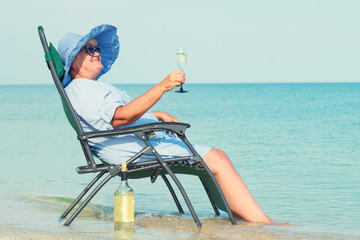 An elderly woman sits on the beach on a chaise longue and drinking wine.	