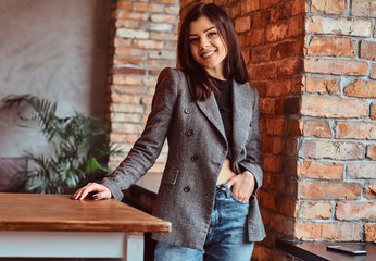 Smiling sensual brunette woman dressed in trendy clothes posing in a room with loft interior, looking at camera.