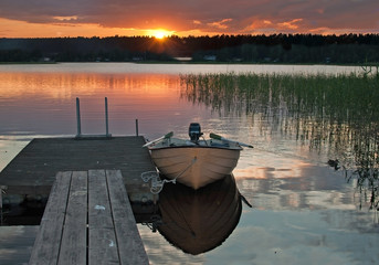 Small boat moored by wooden jetty at peaceful colorful sunset by a lake with sky reflections in tranquil water in Varmland, Sweden.