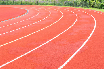 the curve of an empty 6 lane stadium running track bordering a fence with greenery behind it