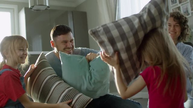 Handheld shot of happy family with two children of elementary school age playing together in living room and having pillow fight, slow motion