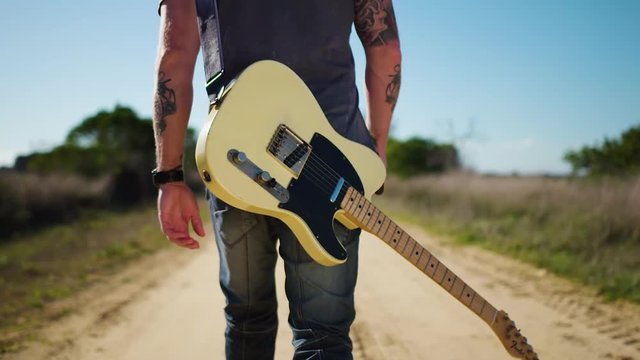 Cool musician walking down dusty track with guitar.