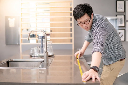 Young Asian man using tape measure for measuring granite countertops on modern kitchen counter in showroom. Shopping furniture for home improvement. Interior design concept