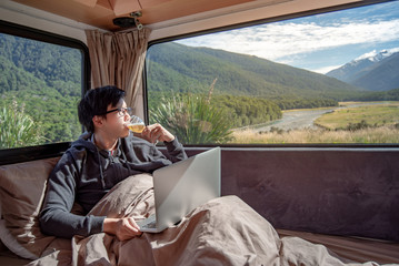 Fototapeta premium Young Asian man drinking beer and working with laptop computer on the bed in camper van with mountain scenic view through the window, digital nomad concept