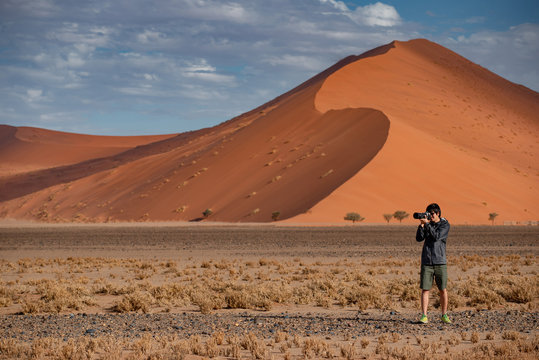 Young male traveler and photographer taking photo in Namib desert with orange sand dune in the background. Travel Namibia, Africa. Photography concept