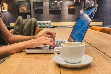 Woman's hand typing in a notebook keyboard with a coffee cup next to it. Wood table, modern design.