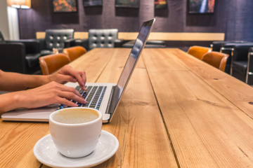 Woman's hand typing in a notebook keyboard with a coffee cup next to it. Wood table, modern design.