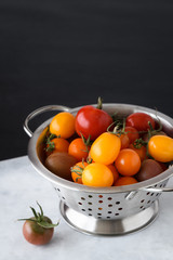 Freshly picked heirloom tomatoes, yellow, red, and purple, in a stainless steel colander on a gray marble slab, black background

