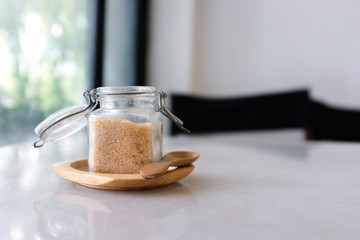 Close up of open small glass jar of brown sugar on wooden tray with wooden spoon on a white marble table with blurred background