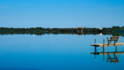 Bemidji, Minnesota, the 2018 Best Town in Minnesota is seen across Lake Irving, the first lake on the Mississippi River, on a sunny day. A dock is seen in the foreground.