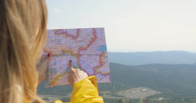 Back view of Caucasian female hiker in yellow raincoat stands in the mountains with a map in hand