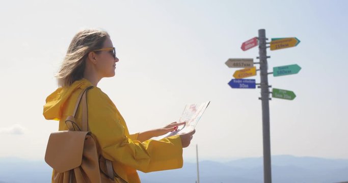 Young female hiking im yellow raincoat with a backpack in mountains holding paper map in hands.