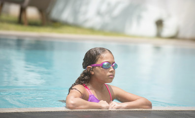 Young child  wearing goggles relaxing at edge of the pool after swimming and playing showing her wet curly hair