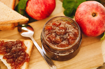 Apple jam on toast and in jar, fresh red apples on a cutting board on a wooden table. Delicious breakfast, rustic style.