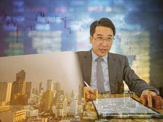 Happy asian business man sitting on his desk with stock market on cityscape background