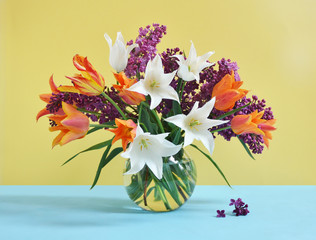 Bouquet of white and orange tulips and lilac in glass vase on yellow background