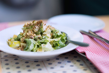 Improvised fresh Caesar salad with green lettuce, walnuts and pear slices on a white plate standing on a table with checkered red and white napkin