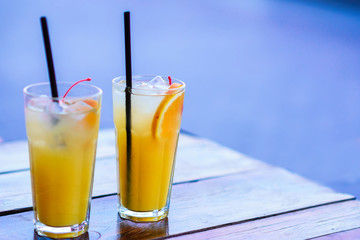 Two high glasses of cold orange fresh lemonade with ice cubes, cherry and coctail straws standing on a lacquered wooden table in the evening.