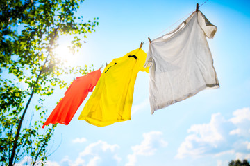 Photo of three different colored T-shirts hanging on ropes against blue sky with tree tops.