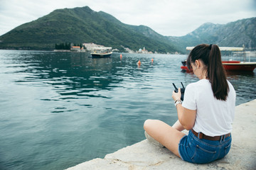 young adult woman sitting on pier with beautiful view of sea nd mountains. drone controller