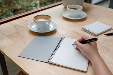 Closeup image of a hand writing down on a white blank notebook with coffee cup on wooden table