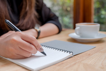 Closeup image of a woman's hand writing down on a white blank notebook with coffee cup on wooden table