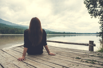 Closeup image of an asian woman sitting alone on an old wooden pier by the river with sky and mountain background