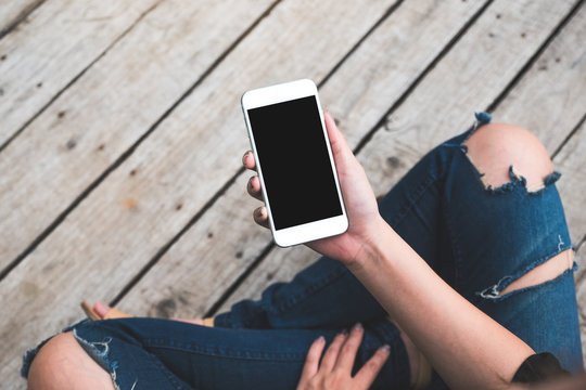 Top view mockup image of a woman holding white mobile phone with blank black screen while sitting on an old wooden floor background
