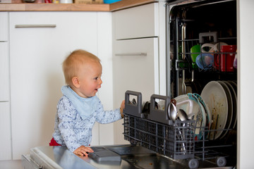 Toddler child, boy, helping mom, putting dirty dishes in dishwasher at home, .