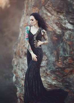 Brunette woman with tattoos posing with a snake. A girl in a black long dress in the desert with a Python.