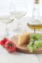 On the table is cheese, tomatoes, grapes, olive oil, parsley, two wineglasses and a bottle of white wine. Also on the white table is a cutting board and a knife.