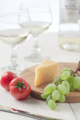 On the table is cheese, tomatoes, grapes, olive oil, parsley, two wineglasses and a bottle of white wine. Also on the white table is a cutting board and a knife.