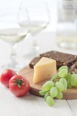 On the table is cheese, tomatoes, grapes, olive oil, parsley, two wineglasses and a bottle of white wine. Also on the white table is a cutting board and a knife.