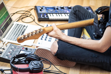 Male guitarist playing electric guitar at modern home studio or rehearsal room. Young man producing music with electronic effects processors, synthesizer and laptop