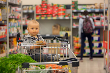 Fototapeta premium Toddler baby boy, sitting in a shopping cart in grocery store, smiling and eating bread