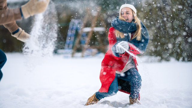 Young Beautiful Couple Throws Snow At Each Other While Snow Falls. Happy Man And Woman Playing With Snow In The Yard Of Their Idyllic House. Family Enjoying Winter.