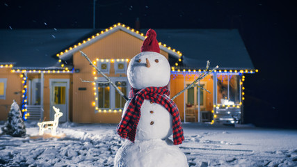 Funny Snowman Wearing Hat and Scarf Standing in the Backyard of the Idyllic House Decorated with Garlands on Christmas Eve. Soft Snow is Falling on that Magical Winter Evening.