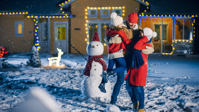 Happy Young Family Portrait In The Falling Snow, Father Embraces Wife And Holds Daughter. Family Enjoying Winter Holiday In The Front Yard Of Their Idyllic House Decorated With Garlands.