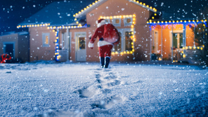 Low Angle Shot of Santa Claus with Red Bag, Walks into Front Yard of the Idyllic House Decorated...