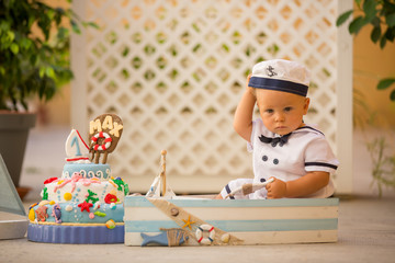 Little toddler boy, dressed as sailor, celebrating his first birthday with sea theme cake, boat,...