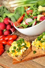 Close-up of wholemeal bread with scrambled eggs, fresh herbs, spring onions, tomatoes and salad bowl in background - healthy breakfast - vertical photo