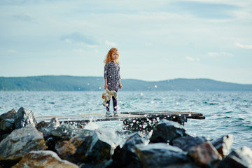 A sad child with a toy stands on the pier by the sea and looks a